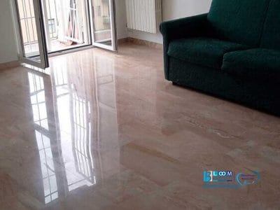 Una stanza per lo più vuota con un pavimento in marmo lucido, un divano verde, un termosifone bianco e porte a vetri aperte che conducono a un balcone con vista sugli edifici cittadini, il tutto mantenuto in modo impeccabile dall'impresa di pulizie De Amicis Milano.