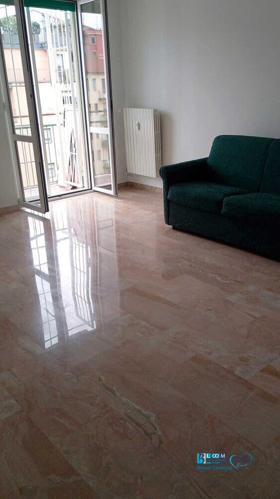 Una stanza per lo più vuota con un pavimento in marmo lucido, un divano verde, un termosifone bianco e porte a vetri aperte che conducono a un balcone con vista sugli edifici cittadini, il tutto mantenuto in modo impeccabile dall'impresa di pulizie De Amicis Milano.