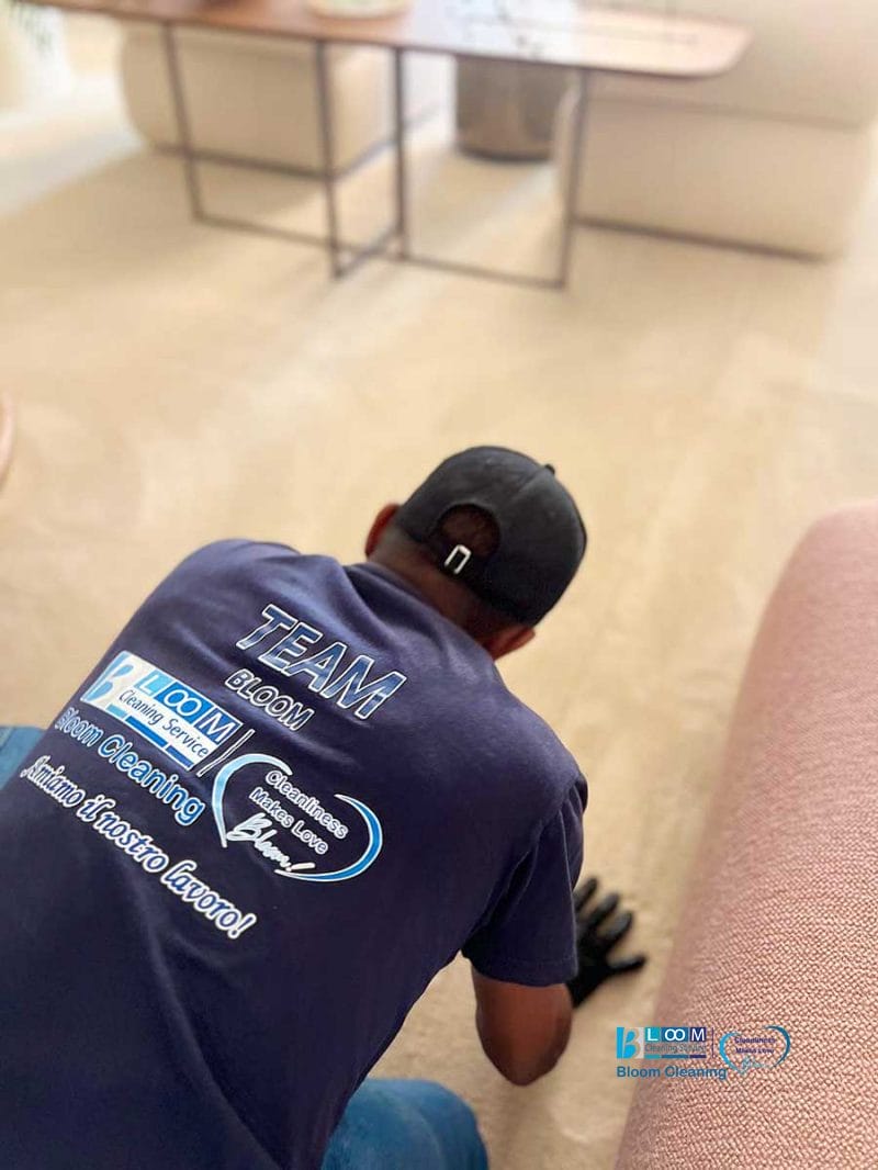 Persona con maglietta e berretto blu della squadra che pulisce un pavimento in moquette accanto a un divano rosa, specialista nella pulizia della moquette zona Amendola.