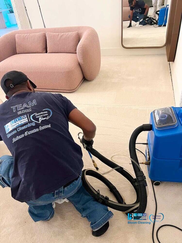 Uomo che pulisce un tappeto chiaro con una macchina blu per la pulizia di tappeti vicino a un divano rosa e un grande specchio a parete, offrendo servizi di pulizia moquette Milano.