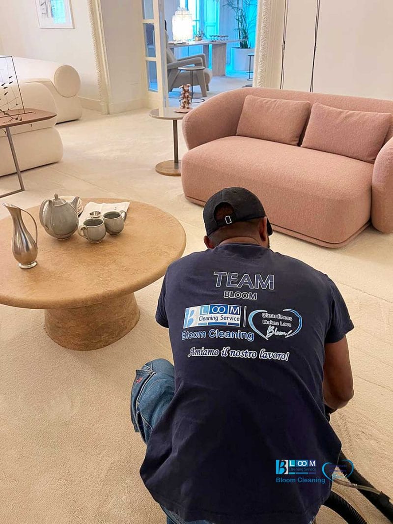 Una persona con una maglietta "Team Bloom" pulisce un moderno soggiorno beige con un tavolino rotondo e un divano, specializzata nella pulizia moquette Milano per tappeti impeccabili.