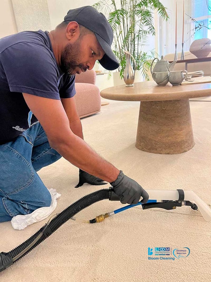 Un uomo con i guanti pulisce un tappeto beige con un aspirapolvere in un soggiorno moderno, dimostrando che la pulizia professionale della moquette zona Amendola garantisce risultati impeccabili.