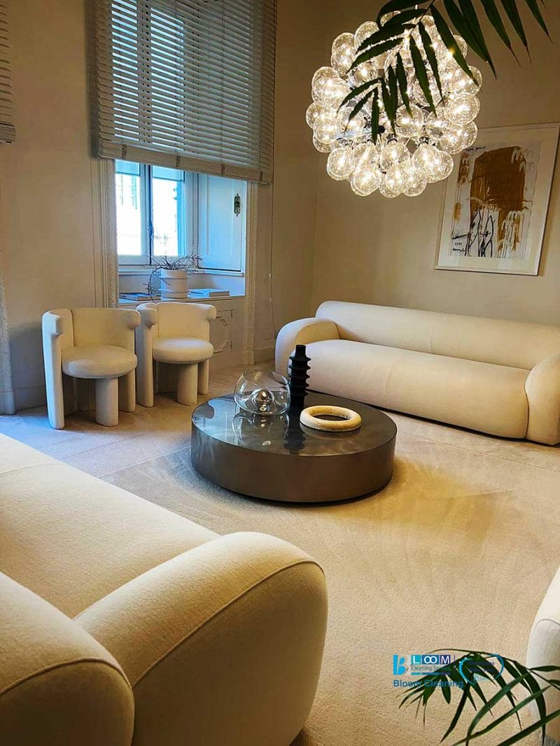 Soggiorno moderno con divani color crema, tavolino rotondo, lampadario scultoreo e ampia finestra con tende: ideale per chi desidera mantenere l'ambiente fresco e invitante.