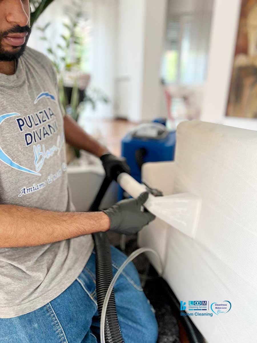 Un professionista esegue il lavaggio divani domicilio Monza, pulendo un divano bianco con un aspirapolvere per tappezzeria, indossando guanti e una maglietta grigia firmata.