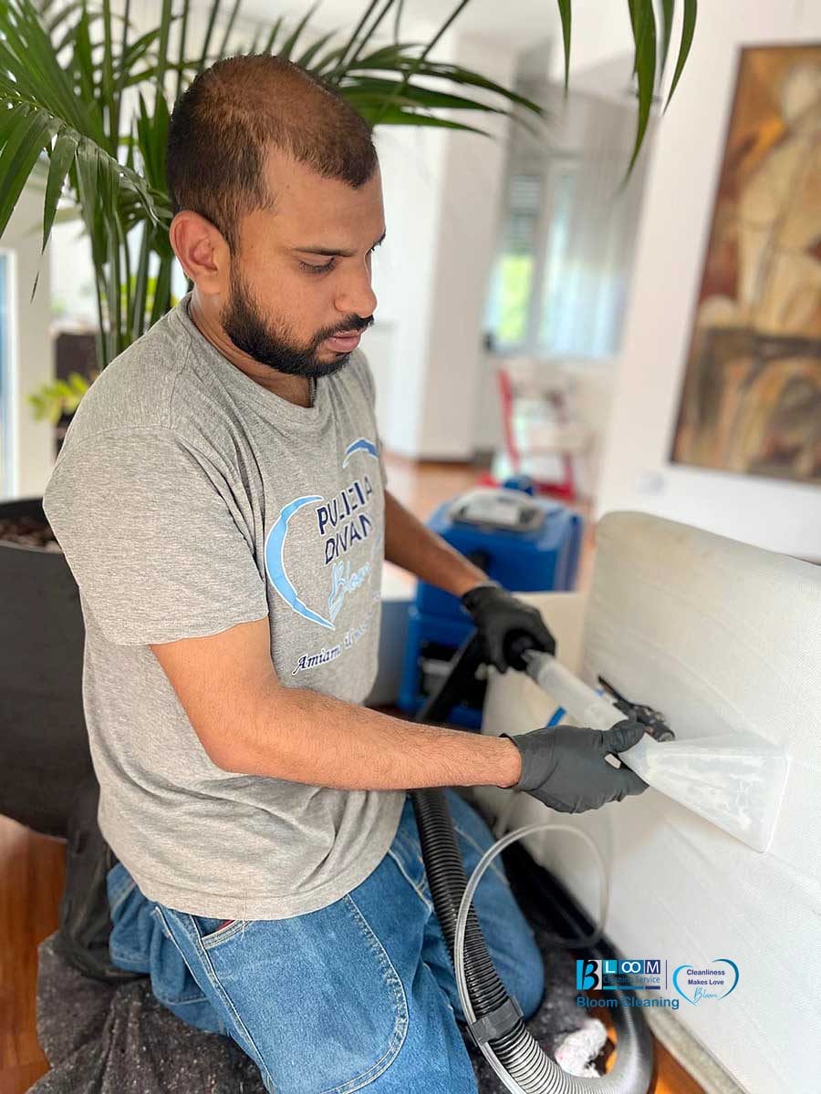Uomo che pulisce un divano bianco con un aspirapolvere, indossando guanti e una maglietta grigia, in un ambiente interno: servizio professionale di lavaggio divani domicilio Monza.