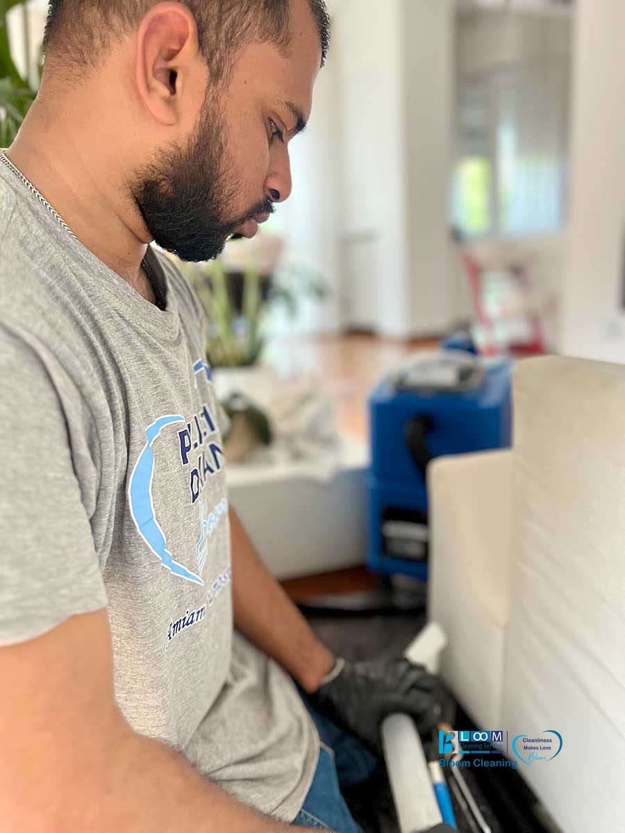 Un uomo con una camicia grigia e guanti neri pulisce un divano bianco con uno strumento di pulizia, dimostrando i servizi professionali di lavaggio divani domicilio Monza all'interno di un'abitazione.