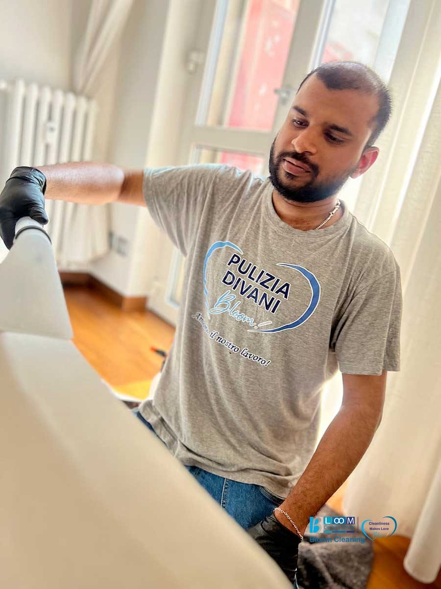 Un uomo che indossa i guanti pulisce una superficie bianca in un ambiente interno, indossando una maglietta con la scritta "Pulizia Divani Bloom", e offre i suoi servizi di lavaggio divani a domicilio a Monza.