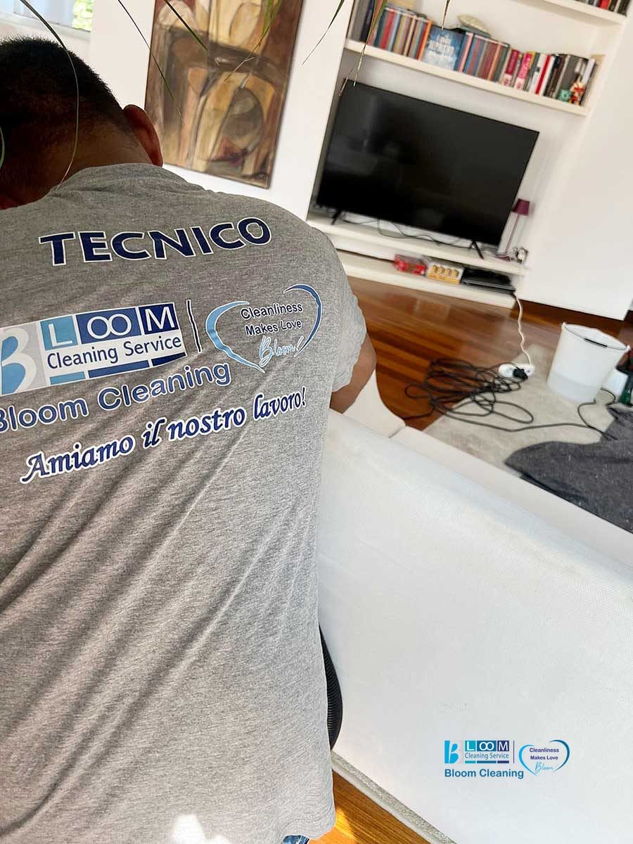 Un tecnico con la ditta "Bloom Cleaning Service" esegue un lavaggio divani domicilio Monza vicino a una TV e a cavi aggrovigliati in un soggiorno moderno.