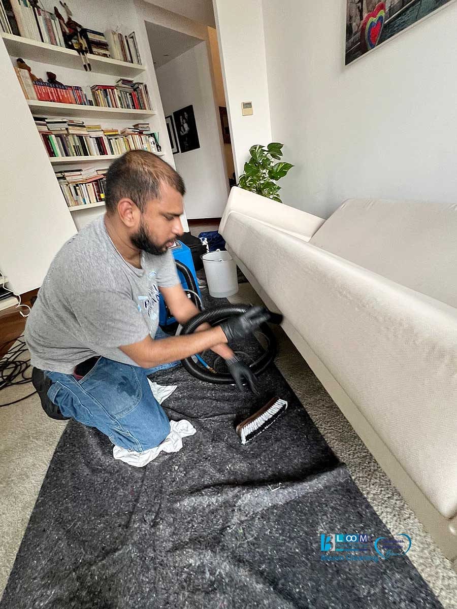 Un uomo che indossa i guanti pulisce un divano bianco in un soggiorno con librerie sullo sfondo, che mette in mostra il lavaggio divani domicilio Monza per risultati impeccabili direttamente a casa tua.