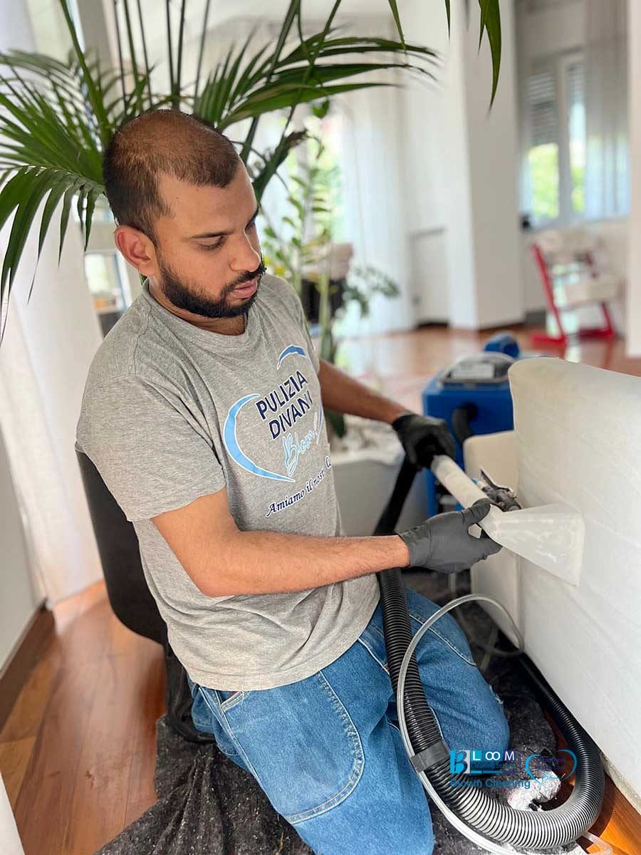 Uomo che pulisce un divano bianco con l'aspiratore, indossa guanti e una maglietta "Pulizia Divani". Servizio professionale di lavaggio divani domicilio Monza.
