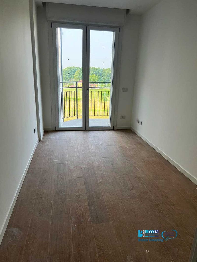 Stanza vuota con pavimento in legno, pareti bianche e una porta a vetri che conduce a un balcone con vista sul verde, perfettamente rinfrescata dopo la pulizia post cantiere Nerviano.