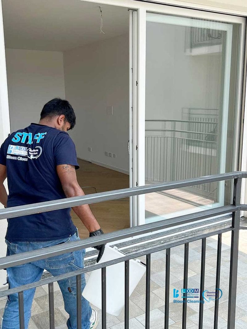 Un uomo che indossa una maglietta con la scritta "STAFF" sposta un oggetto di grandi dimensioni attraverso una porta scorrevole in vetro su un balcone durante la pulizia post cantiere Nerviano.