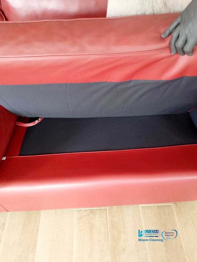 Una mano solleva un cuscino rosso del divano, rivelando lo spazio di archiviazione vuoto foderato di nero sottostante, ideale per organizzare durante la pulizia degli appartamenti Bollate.