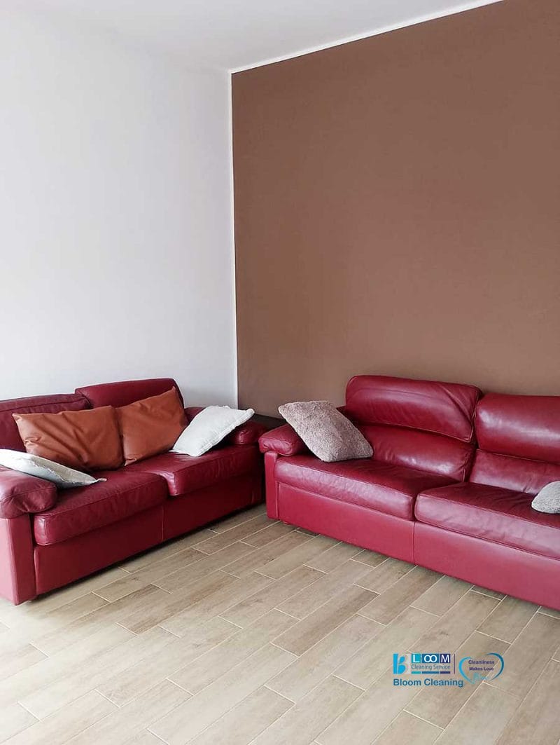 Due divani in pelle rossa con cuscini si trovano in un angolo di una stanza con una parete decorativa marrone e un pavimento in legno, a dimostrazione del comfort che ci si può aspettare dopo la pulizia appartamenti Bollate.