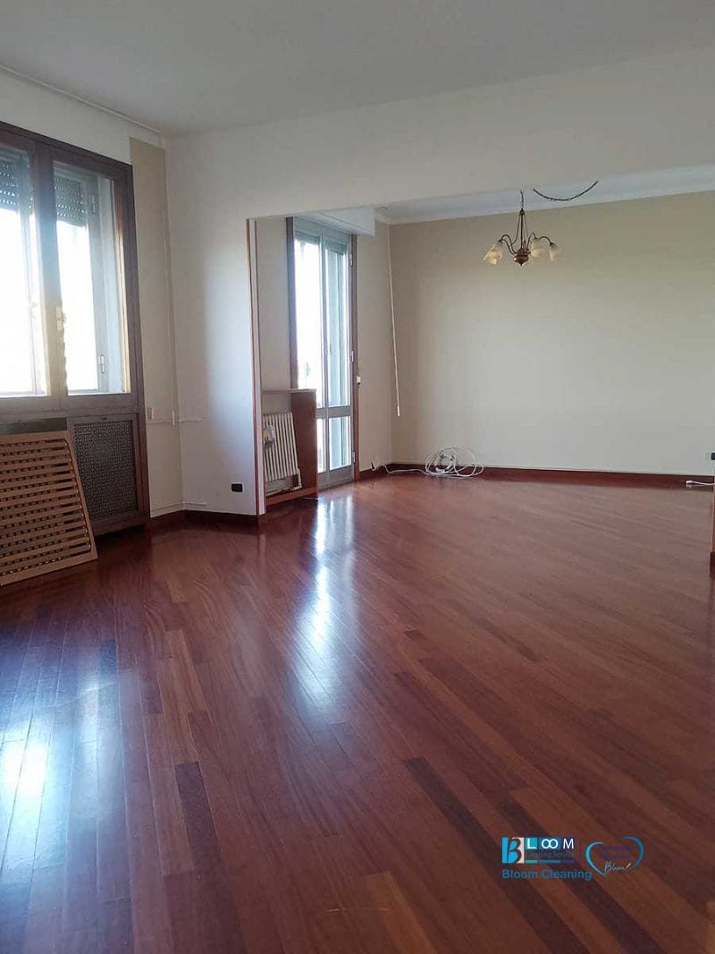 Stanza vuota con pavimento in legno, ampie finestre, pareti beige e una lampada a soffitto appesa al soffitto: ideale per un nuovo inizio dopo la pulizia dell'appartamento Novate Milanese.