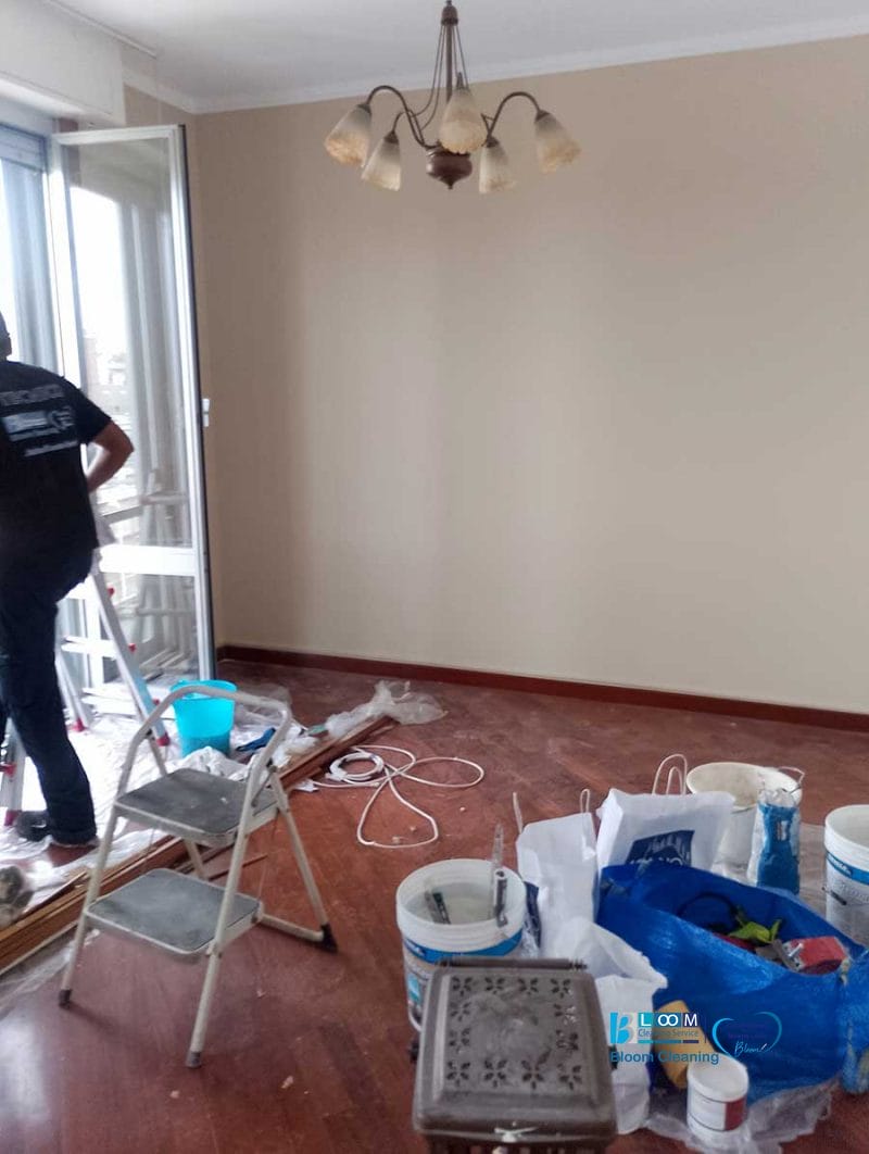 Una stanza in fase di ristrutturazione con materiali per la pittura e una scala a pioli; una persona è in piedi vicino a una porta-finestra aperta, preparandosi per la pulizia dell'appartamento Novate Milanese.