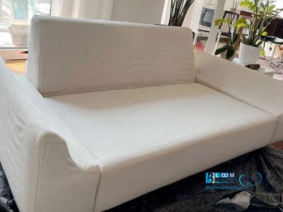 Divano bianco moderno dalle linee pulite in un soggiorno luminoso, vicino a piante da interno e ampie finestre: perfetto per chi cerca un lavaggio divani domicilio Monza per mantenere il proprio spazio fresco ed elegante.