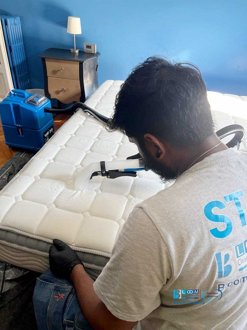 Uomo che pulisce un materasso con una macchina a vapore in una camera da letto, indossando guanti e una camicia grigio chiaro, assicurando igiene profonda con lavaggio materassi a domicilio Novara.