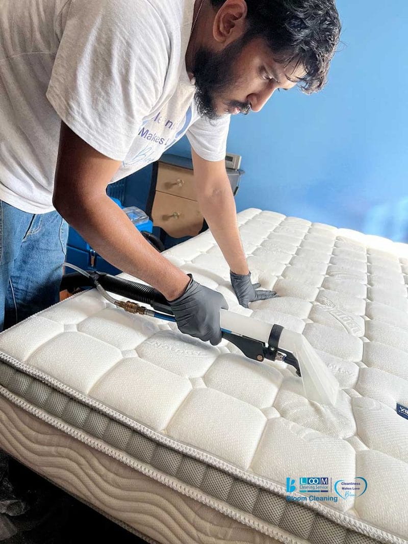 Uomo con guanti che pulisce un materasso con l'aspirapolvere in una camera da letto dalle pareti blu, assicurando igiene profonda. Lavaggio professionale materassi a domicilio disponibile a Novara per una biancheria da letto immacolata e rinfrescata.