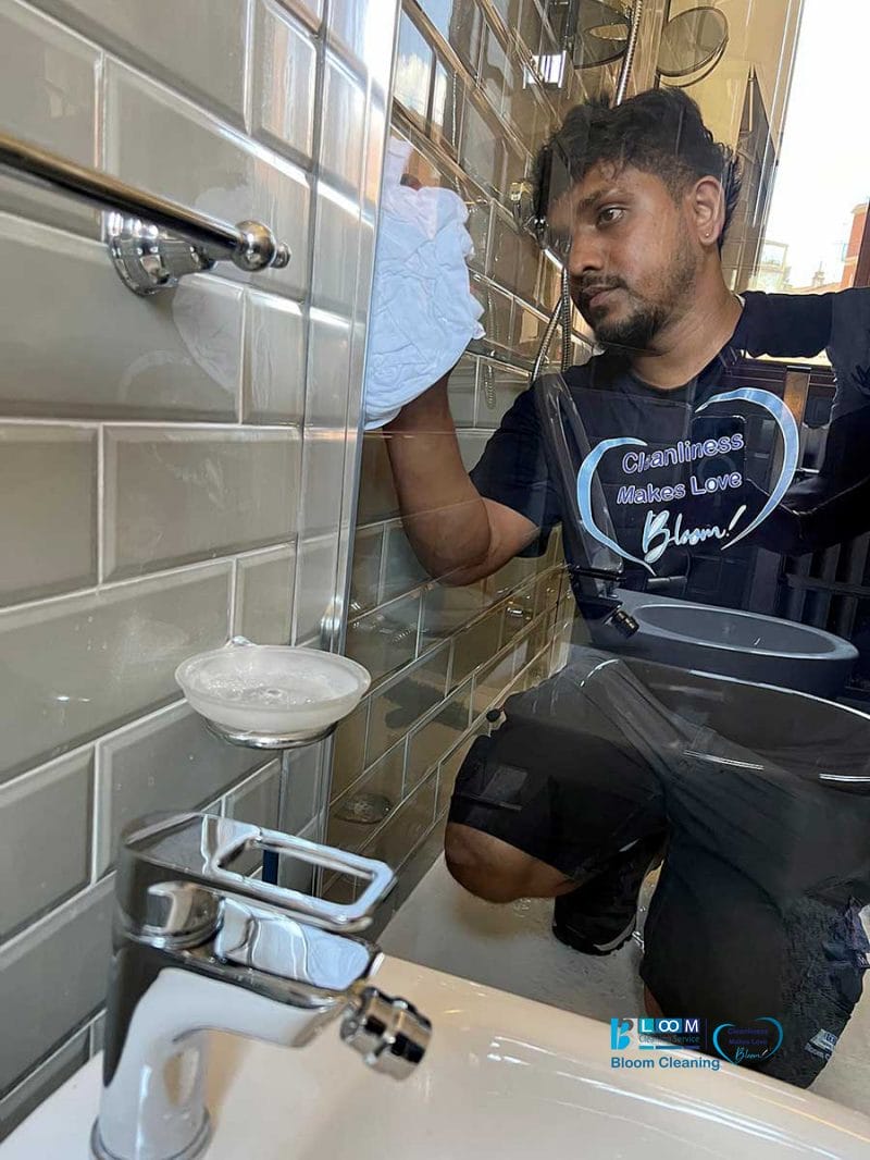 Un uomo con la maglietta "Cleanliness Means Love" pulisce lo specchio del bagno con un panno dopo la pulizia post-ristrutturazione in zona Solari a Milano.