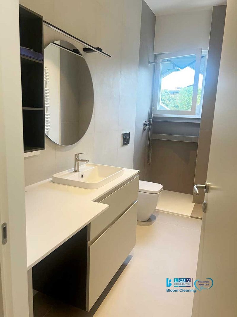 Bagno moderno con specchio rotondo, lavandino bianco e toilette. Un'ampia finestra riempie lo spazio di luce naturale, perfetta dopo una pulizia post ristrutturazione Milano 2 per una sensazione di pulizia e freschezza.