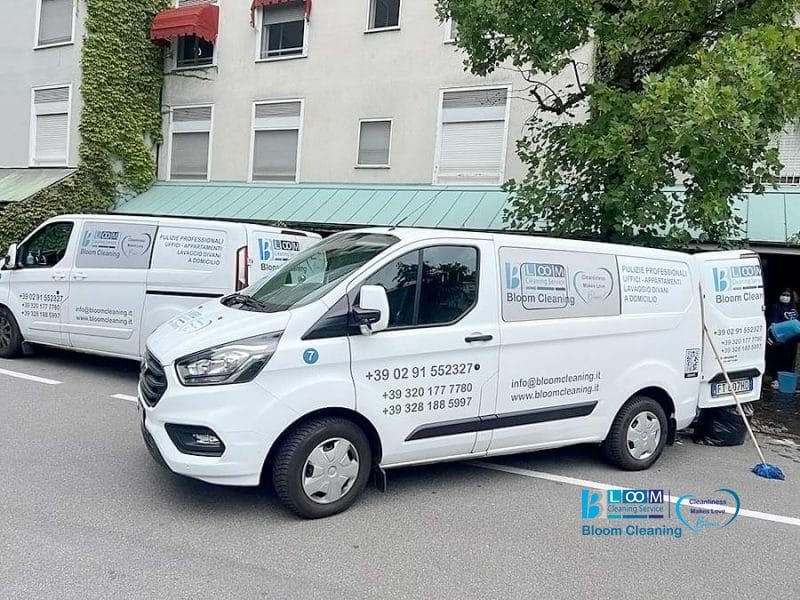 Due furgoni bianchi del servizio Bloom Cleaning, che rappresentano i professionisti della pulizia di Milano, sono parcheggiati in una strada vicino a un edificio con del verde.