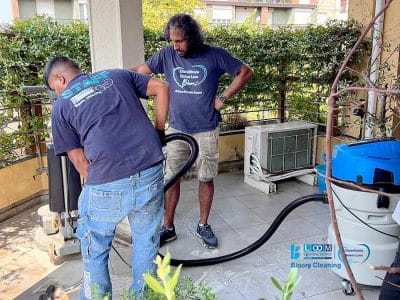 Due uomini eseguono la pulizia profonda in un patio piastrellato di un appartamento, con piante e un condizionatore nelle vicinanze.