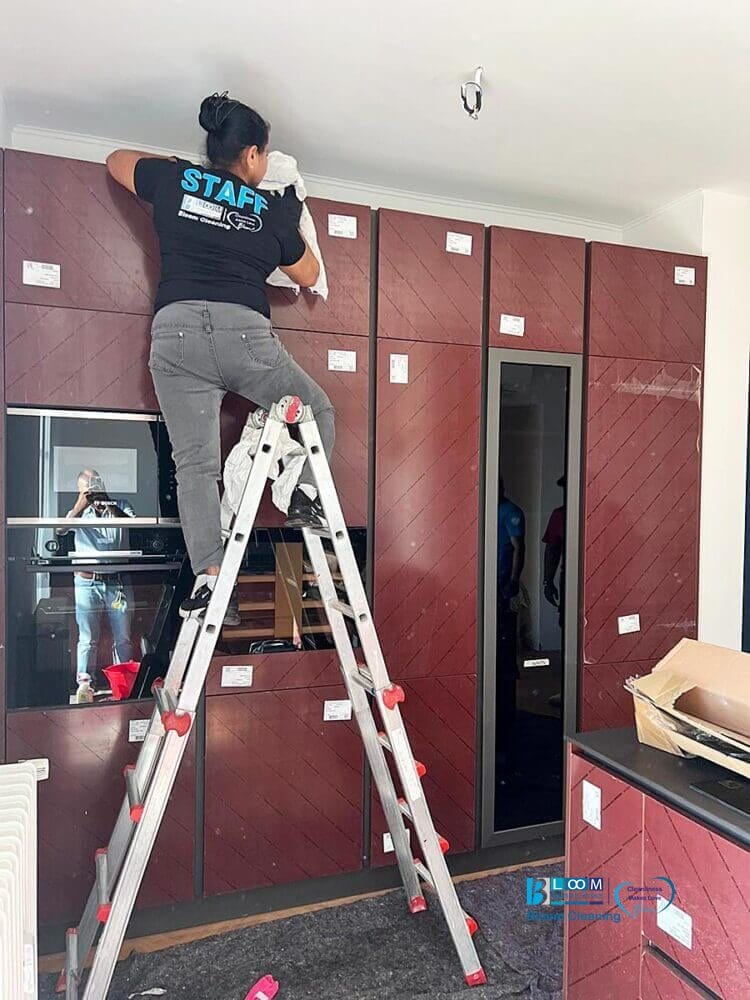Durante le pulizie finali cantiere in appartamenti nuovi, una persona su una scala pulisce i mobili alti della cucina con le etichette; un'altra persona è visibile nel riflesso.