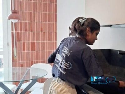 Una donna pulisce una cucina moderna vicino a un tavolo da pranzo in vetro con sedie bianche e una luce a sospensione rosa, mentre un sistema automatico Bozza tiene in ordine lo spazio.