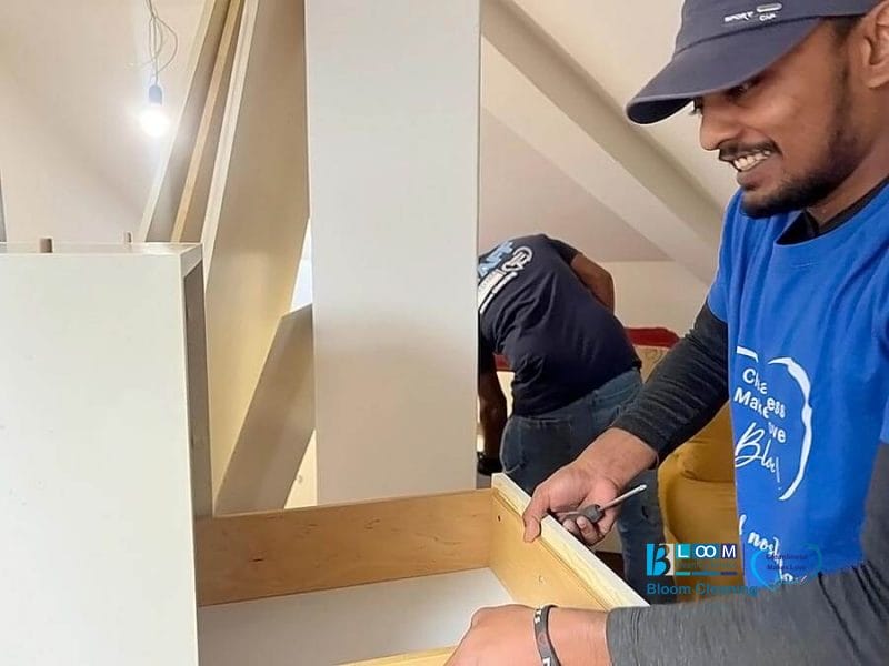 Due persone che assemblano mobili in legno in una stanza luminosa; una persona in primo piano sorride tenendo in mano un pezzo di legno e un cacciavite, indossando una camicia blu e un berretto, ideale per chi cerca servizio sgombero e pulizia casa Milano.