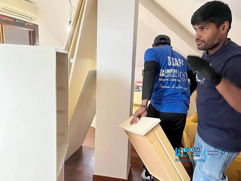 Due traslocatori, parte di un team di Servizio Sgombero e Pulizia Casa Milano, montano o smontano mobili bianchi in una stanza con pavimenti in legno e un divano giallo. Uno porta un pannello mentre l'altro, che indossa una maglietta dello