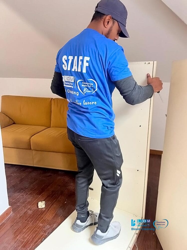 Una persona con maglietta e berretto blu "STAFF" monta i mobili in una stanza con pavimento in legno e divano giallo, mostrando il servizio sgombero e pulizia casa Milano per uno spazio abitativo rinnovato.