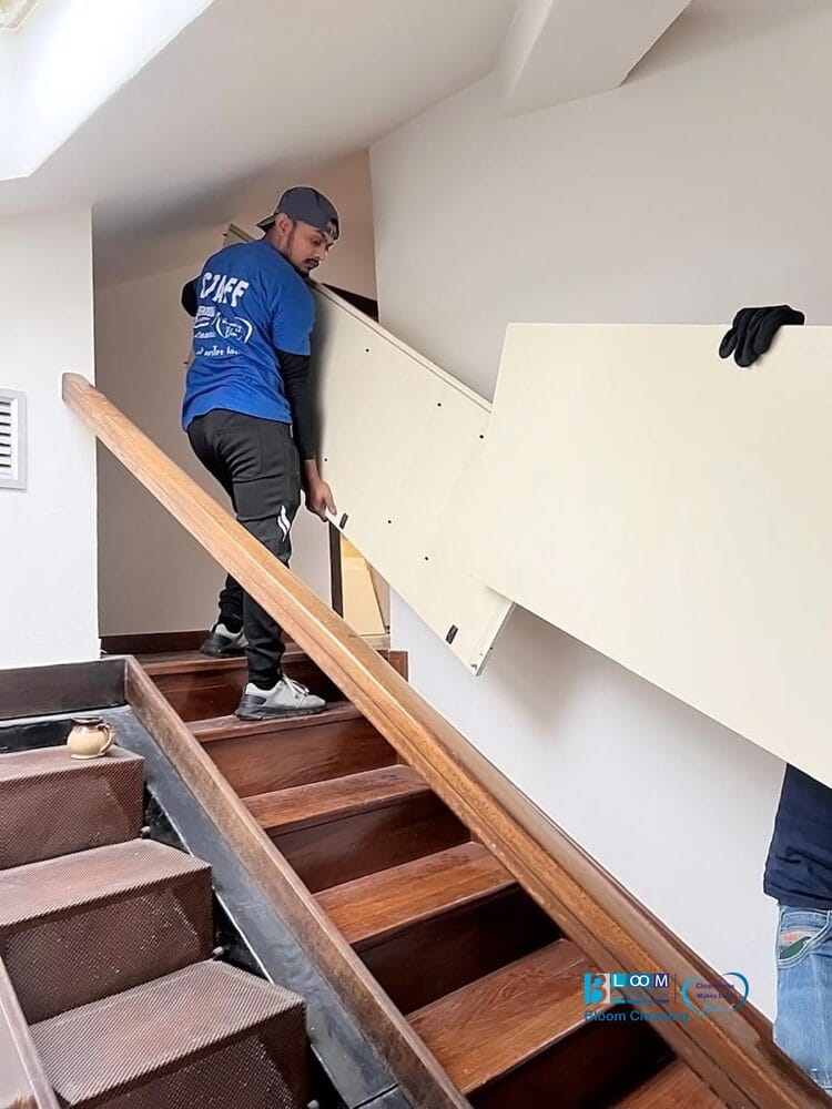 Due persone stanno manovrando un grande pannello rettangolare su una stretta scala di legno, parte di un servizio sgombero e pulizia casa Milano, una in camicia blu e berretto, l'altra visibile solo da una mano guantata.
