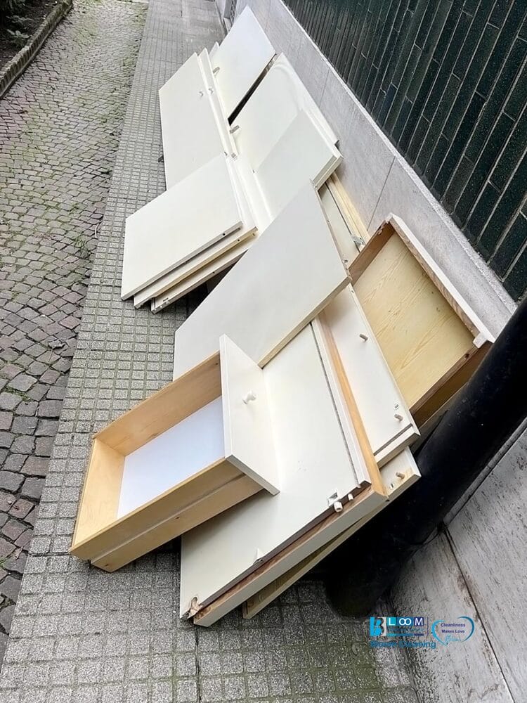 Mobili smontati in legno bianco, compresi cassetti e pannelli, sono accatastati su un marciapiede accanto a un tombino e a un muro di piastrelle verdi, apparentemente in attesa dello smaltimento mobili ingombranti Milano o del servizio sgombero e pulizia casa Milano.