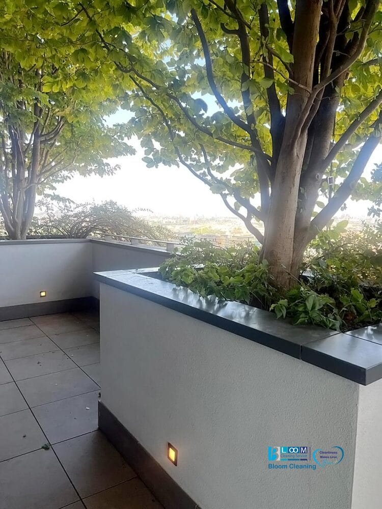 Una moderna terrazza esterna con grandi alberi in vaso, fronde verdi e pavimento in piastrelle, ideale dopo una pulizia post sgombero appartamento Milano. Le luci gialle a parete illuminano le pareti bianche delle fioriere in un paesaggio urbano.