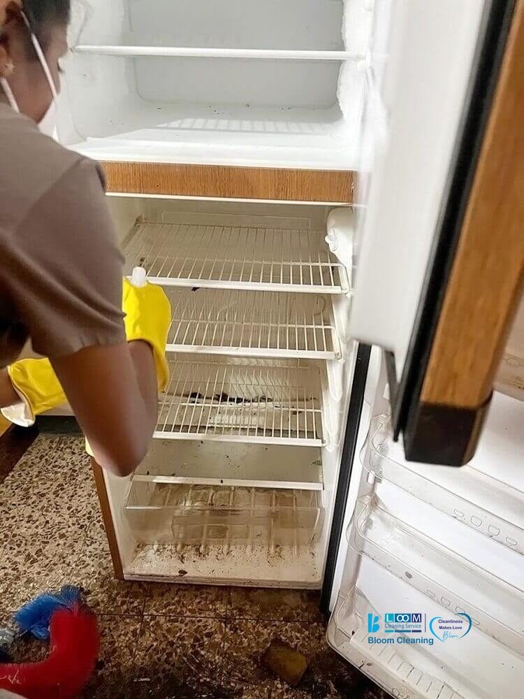 Una persona che indossa guanti gialli pulisce un frigorifero sporco e vuoto con le porte aperte: la sporcizia ricopre i ripiani e il pavimento e i prodotti per la pulizia dell'appartamento di Monza sono sparsi a terra nelle vicinanze.