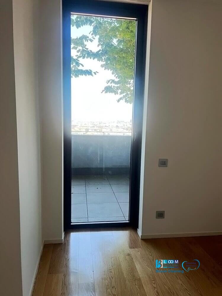 Una porta a vetri in una stanza dalle pareti bianche si apre su un balcone piastrellato con rami di alberi e un paesaggio urbano lontano. La luce del sole si diffonde sul pavimento in legno, evocando la freschezza di una pulizia post sgombero appartamento Milano.