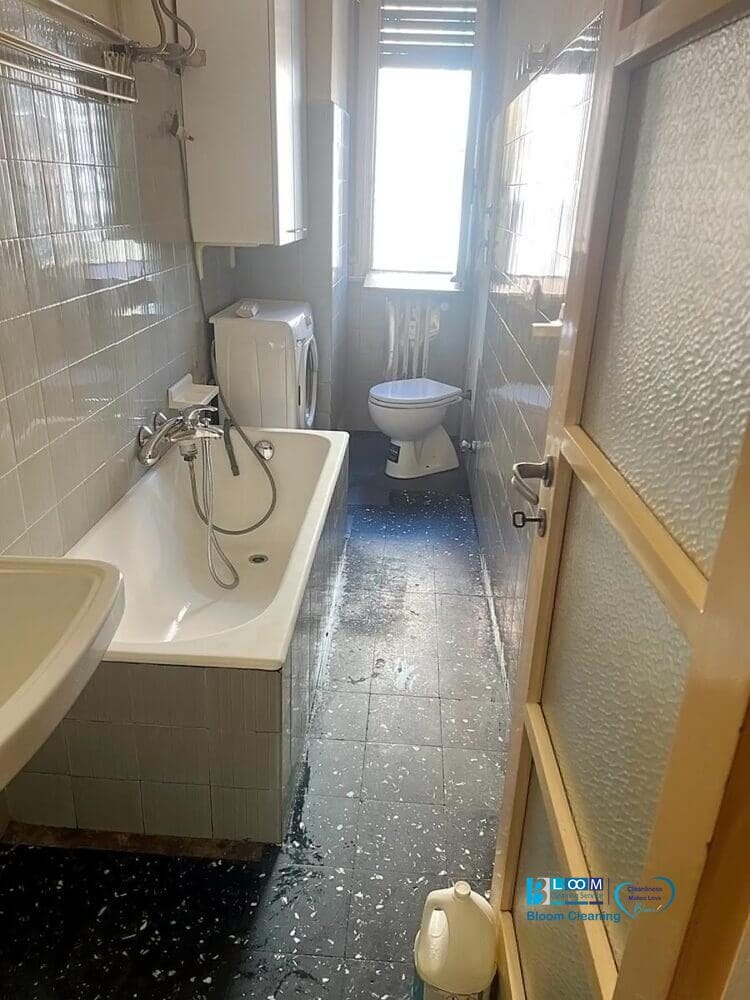 Uno stretto bagno con vasca, lavandino, lavatrice e toilette presenta pareti piastrellate grigie e un pavimento a chiazze scure. La luce naturale entra da una finestra in fondo, ideale per pulire facilmente un appartamento a Monza.