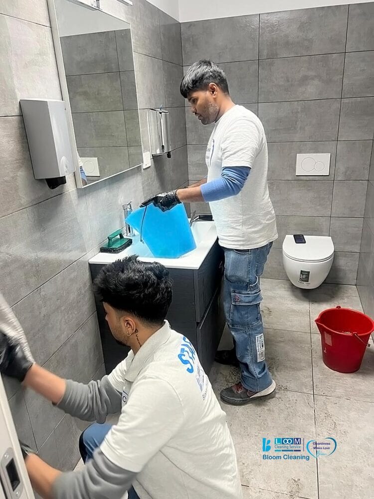 Due persone in camicia bianca e guanti puliscono un bagno moderno in perfetto stile Bozza automatica: una lava il lavandino con un secchio blu, l'altra pulisce una parete. Un secchio rosso è appoggiato sul pavimento vicino al water.