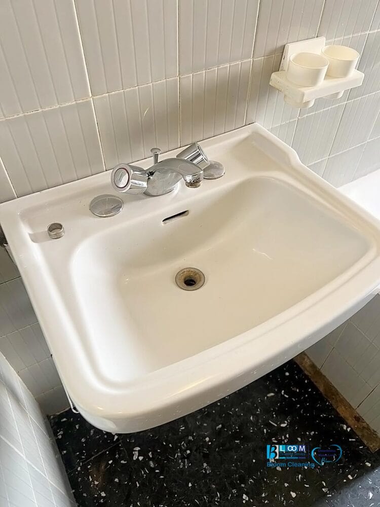 Lavandino da bagno bianco con rubinetto cromato, montato su una parete piastrellata. Sopra il lavabo si trova un portasapone che contiene due piccole tazze bianche. Bozza automatica assicura che il piano di lavoro e l'area circostante rimangano puliti e ordinati.