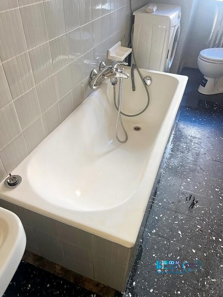 Una vasca da bagno bianca con soffione e rubinetto si trova accanto a una lavatrice in un bagno piastrellato. Sulla lavatrice è riportata la scritta Bozza automatica. Schizzi d'acqua segnano il pavimento a chiazze scure vicino alla vasca, con un gabinetto visibile sullo sfondo.