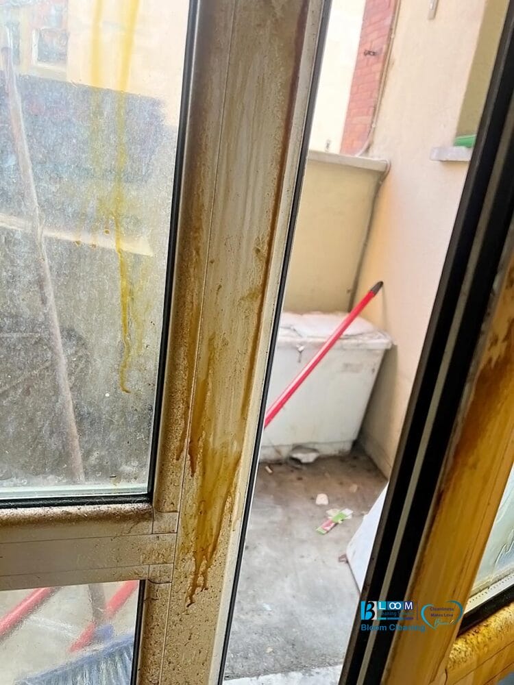 Una porta a vetri con macchie giallo-marroni corre lungo il telaio e conduce a un piccolo balcone sporco, dove una scopa rossa e un contenitore bianco sono appoggiati al muro, quasi fossero stati abbandonati in un momento di automatismo di Bozza.