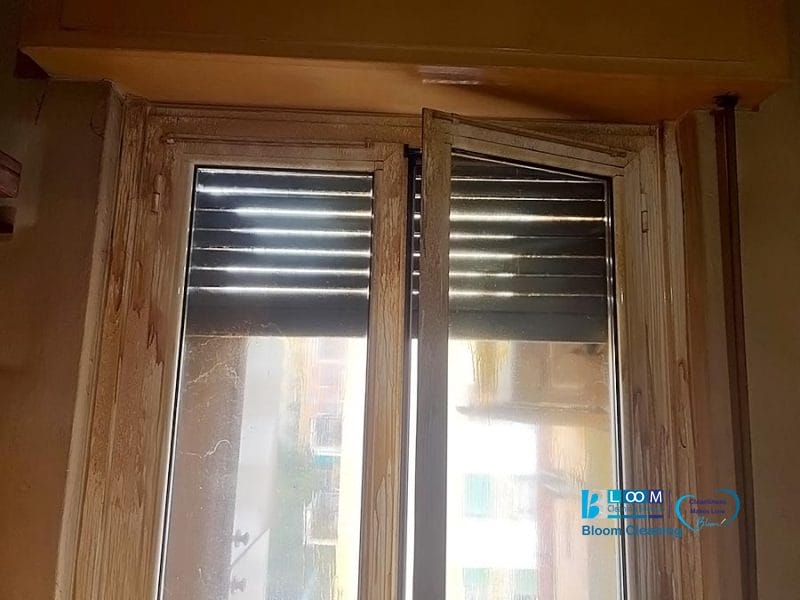 Una finestra con struttura in legno è parzialmente aperta, con tende orizzontali nere parzialmente abbassate in stile bozza automatica. La luce entra dalla finestra e i riflessi di un edificio e del cielo sono visibili sul telaio di vetro usurato.