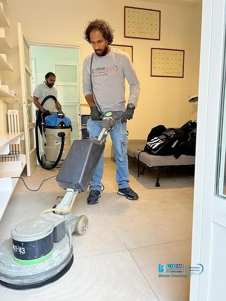 Due uomini puliscono una stanza; uno usa una macchina automatica Bozza per la pulizia dei pavimenti sulle piastrelle, mentre l'altro aziona un aspirapolvere sullo sfondo. La stanza presenta scaffali, oggetti incorniciati alle pareti e un letto con borse.