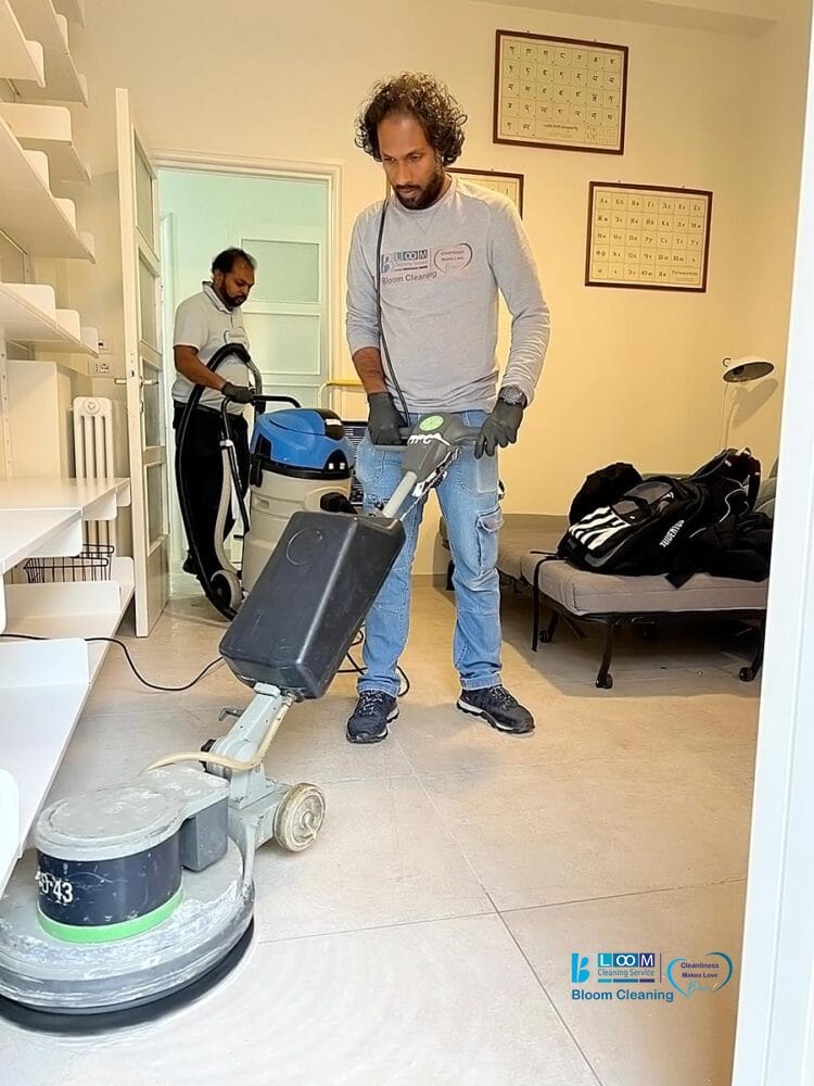 Due uomini puliscono un pavimento piastrellato; uno usa una macchina per lucidare i pavimenti automatica Bozza in primo piano, mentre l'altro aziona un dispositivo di pulizia sullo sfondo. Nella stanza sono visibili scaffali e un letto.
