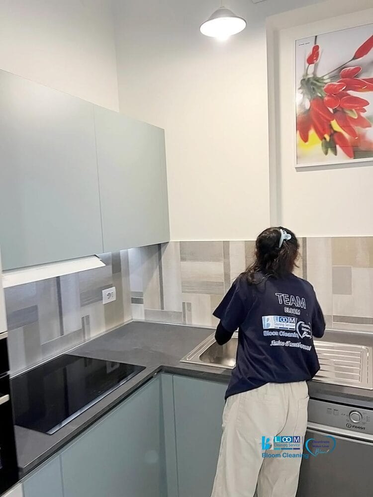 Una persona rivolta verso l'esterno sta pulendo il lavello di una cucina moderna in uno spazio elegante. La cucina, caratterizzata da armadietti chiari e da un backsplash geometrico, reca la scritta 