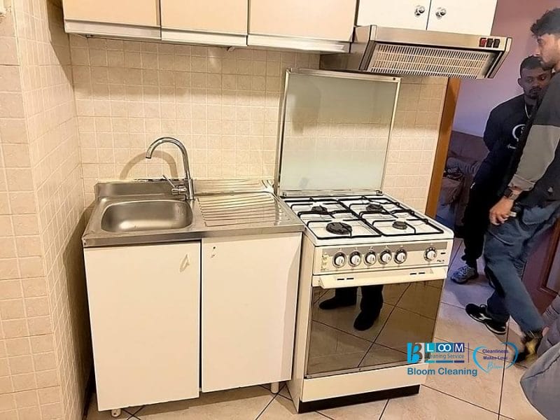 Una piccola cucina con un lavello in acciaio inox, un mobile bianco, un fornello a gas con forno e una cappa aspirante. Sul bancone è presente una Bozza automatica, mentre sullo sfondo sono parzialmente visibili tre persone vicino a una porta.