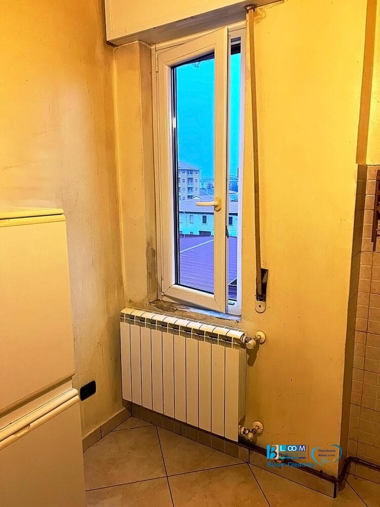 Un piccolo angolo di una stanza dipinta di giallo con un radiatore bianco sotto una finestra. All'esterno sono visibili degli edifici. Si vedono anche parte di un frigorifero e una parete piastrellata. Lo spazio ha un aspetto pulito ma un po' logoro, come una bozza automatica in corso.