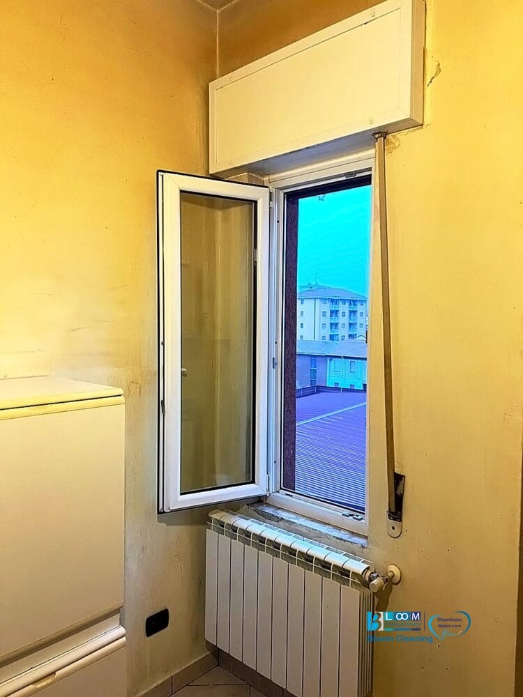 Una piccola stanza dalle pareti gialle presenta una finestra aperta che rivela gli edifici esterni. Sotto la finestra si trova un termosifone bianco, mentre sulla sinistra è visibile parte di un elettrodomestico bianco, forse un frigorifero con bozza automatica.