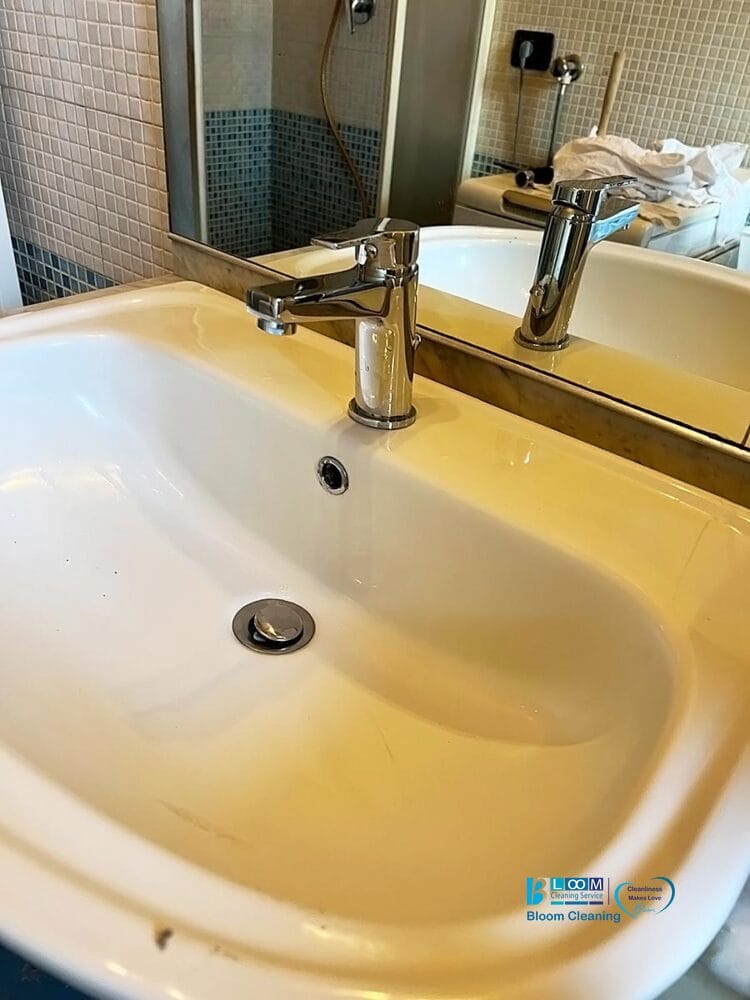 Davanti a uno specchio è raffigurato il lavandino di un bagno bianco con rubinetto argentato. Lo sfondo è caratterizzato da pareti piastrellate beige e blu, alcuni asciugamani sul bancone e una Bozza automatica per una maggiore comodità.
